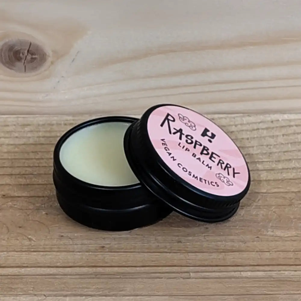 Round black tin labelled in Raspberry Lip Balm, Vegan Cosmetics on a pink label. Container is open with the lid leaning on the bottom revealing the contents.