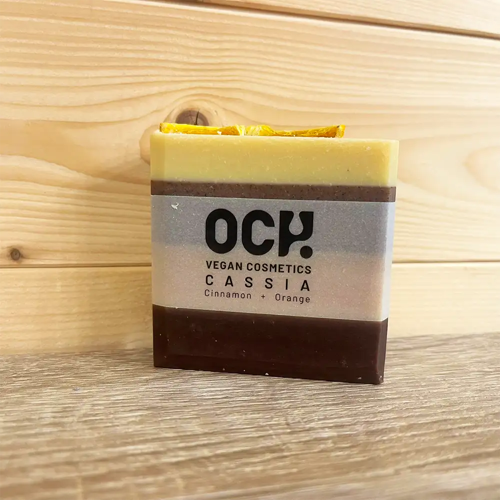Square bar of soap with an opaque band round it with black. Writing states 'Och! vegan cosmetics. Cassia cinnamon and orange'. Soap has four colours in horizontal bands. 