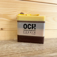 Square bar of soap with an opaque band round it with black. Writing states 'Och! vegan cosmetics. Cassia cinnamon and orange'. Soap has four colours in horizontal bands. 