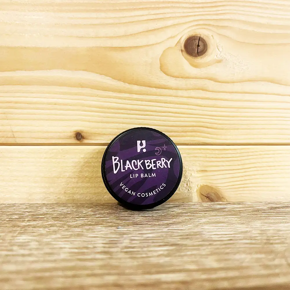 Round tin with a purple front and white writing. White writing states 'Blackberry Lip Balm - Vegan cosmetics'. Tin is on a wooden surface and wooden background. 