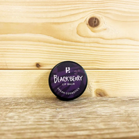 Round tin with a purple front and white writing. White writing states 'Blackberry Lip Balm - Vegan cosmetics'. Tin is on a wooden surface and wooden background. 
