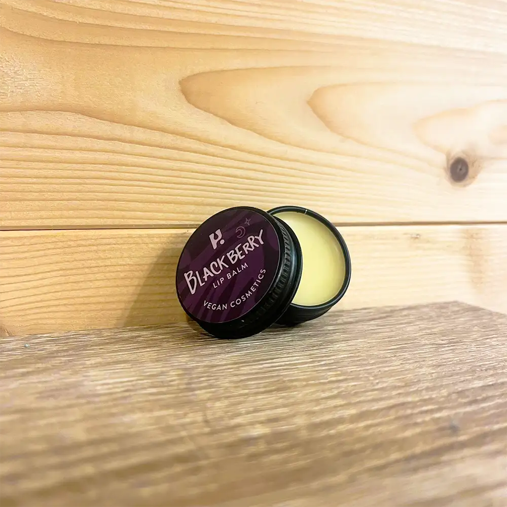 Round tin with a purple front and white writing. White writing states 'Blackberry Lip Balm - Vegan cosmetics'. Tin is open with cream coloured balm visable. Tin is on a wooden surface and wooden background. 