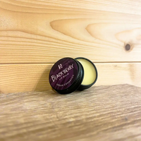 Round tin with a purple front and white writing. White writing states 'Blackberry Lip Balm - Vegan cosmetics'. Tin is open with cream coloured balm visable. Tin is on a wooden surface and wooden background. 