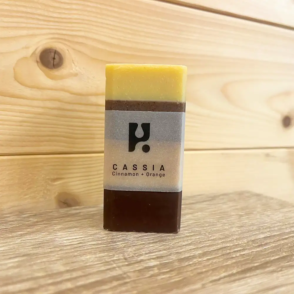 Rectangle bar of soap with an opaque band round it with black. Writing states 'Cassia cinnamon and orange'. Soap has four colours in horizontal bands. 