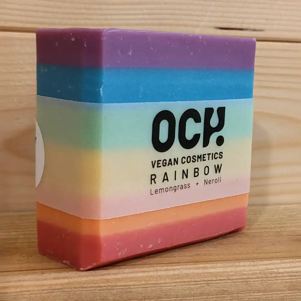 Rainbow coloured soap bar with violet at the top to red at the bottom. Och Vegan Cosmetics - Rainbow - Lemongrass & Neroli on a semi opaque wrapper which covers approximately 50 percent of the height of the bar.