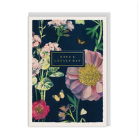 A floral greeting card with a dark navy background, featuring large pink and red flowers, green leaves, butterflies, and a central black label that reads 'HAVE A LOVELY DAY'. The card is shown standing upright against a plain white background.