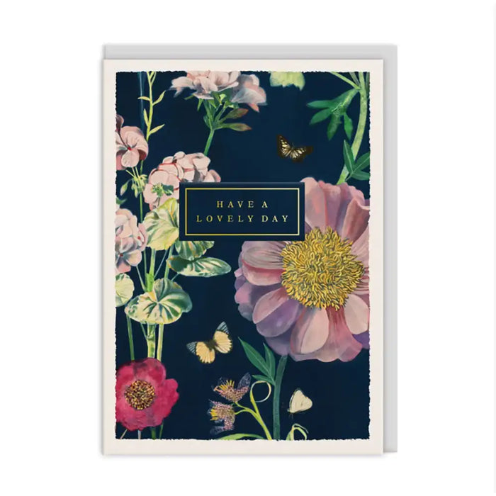 A floral greeting card with a dark navy background, featuring large pink and red flowers, green leaves, butterflies, and a central black label that reads 'HAVE A LOVELY DAY'. The card is shown standing upright against a plain white background.