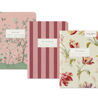 Three Laura Ashley stationery items arranged overlapping on a white background: a floral journal, a pink striped sketchbook, and a floral notebook.