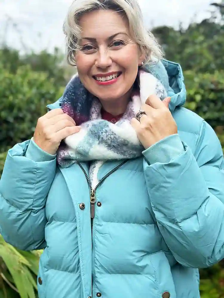 A smiling woman outdoors wearing a light blue puffer jacket and holding a fuzzy multicolor scarf.