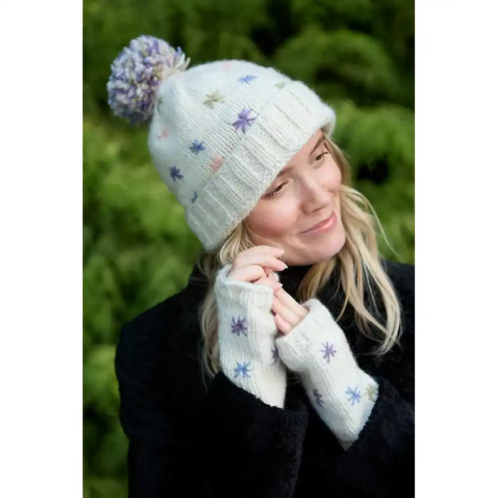 A woman wearing a cream knit beanie with a purple and blue pom-pom and matching knit fingerless gloves, posed outdoors against a soft green background.