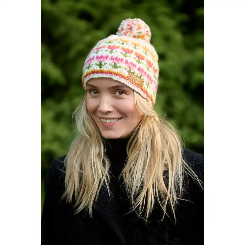 A woman wearing a colorful knit pom-pom beanie hat with multi coloured flowers, photographed outdoors with a blurred green background.