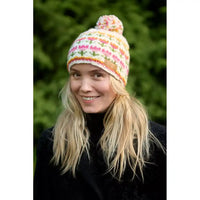 A woman wearing a colorful knit pom-pom beanie hat with multi coloured flowers, photographed outdoors with a blurred green background.