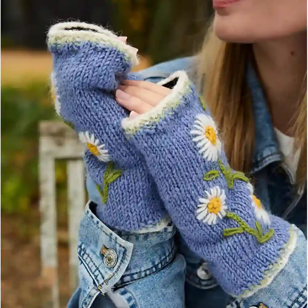 A close-up of a person wearing light blue knit fingerless gloves with white daisy embroidery, yellow centers, and green stems, paired with a denim jacket outdoors.