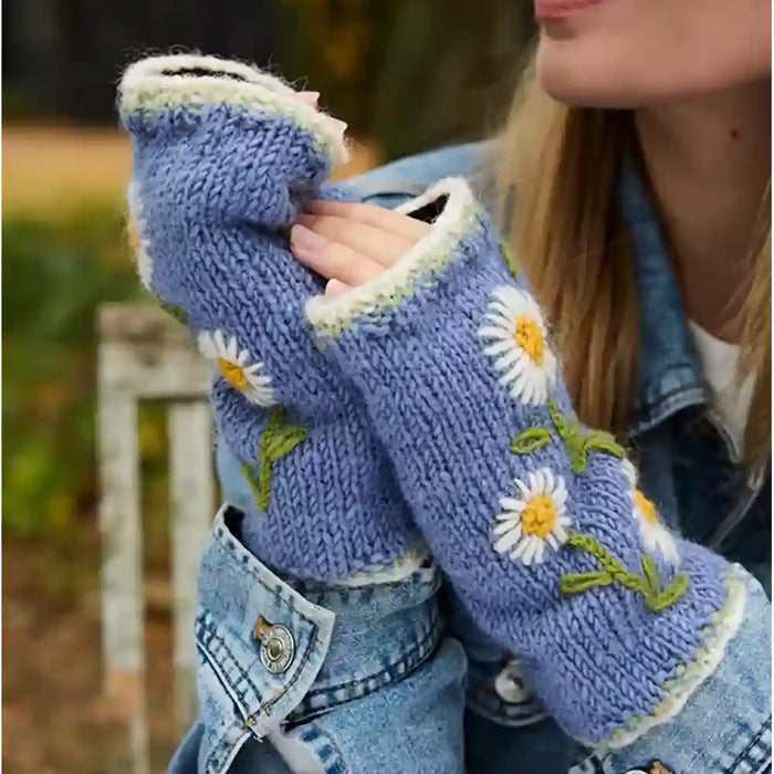 A close-up of a person wearing light blue knit fingerless gloves with white daisy embroidery, yellow centers, and green stems, paired with a denim jacket outdoors.