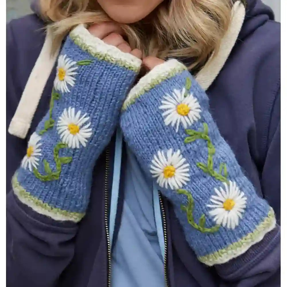 A close-up of a person wearing blue knit fingerless gloves with white daisy embroidery, green stems, and cream trim, paired with a navy hoodie.