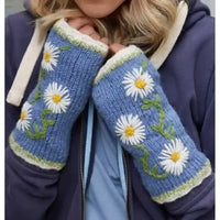 A close-up of a person wearing blue knit fingerless gloves with white daisy embroidery, green stems, and cream trim, paired with a navy hoodie.