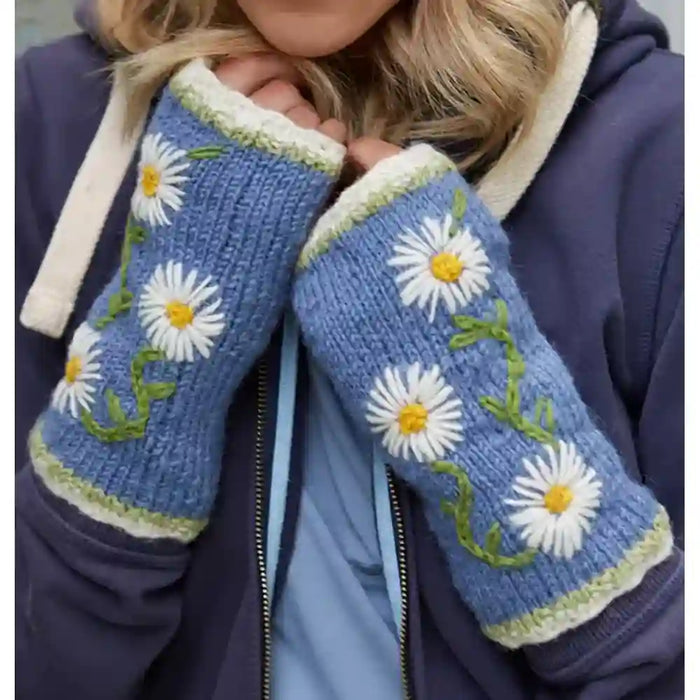 A close-up of a person wearing blue knit fingerless gloves with white daisy embroidery, green stems, and cream trim, paired with a navy hoodie.