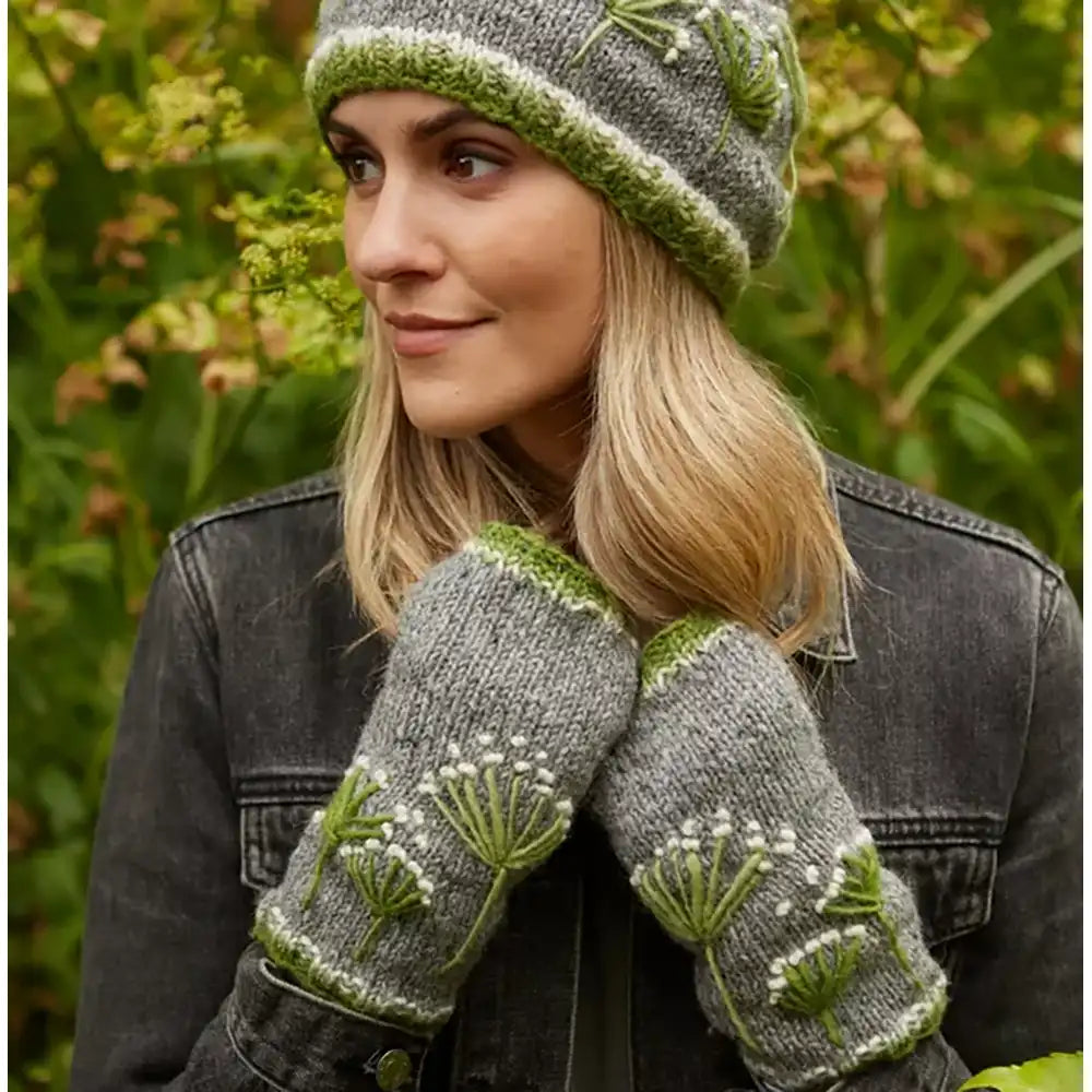 A woman outdoors wearing a gray knit beanie and matching knit fingerless gloves with green and white floral embroidery, paired with a dark denim jacket against a blurred green garden background.