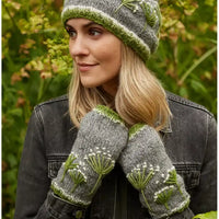 A woman outdoors wearing a gray knit beanie and matching knit fingerless gloves with green and white floral embroidery, paired with a dark denim jacket against a blurred green garden background.