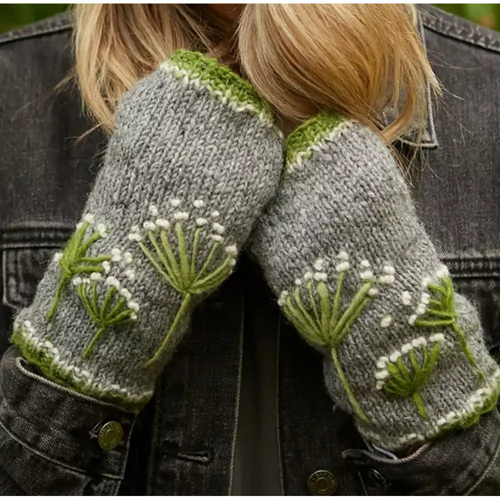 A close-up of a person wearing gray knit fingerless gloves with green and white dandelion embroidery, shown against a dark denim jacket.