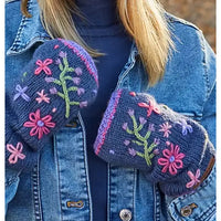 A person wearing a blue denim jacket and a navy knit mitten glove with colorful embroidered flowers and green stems, holding the other mitten in front of the chest.