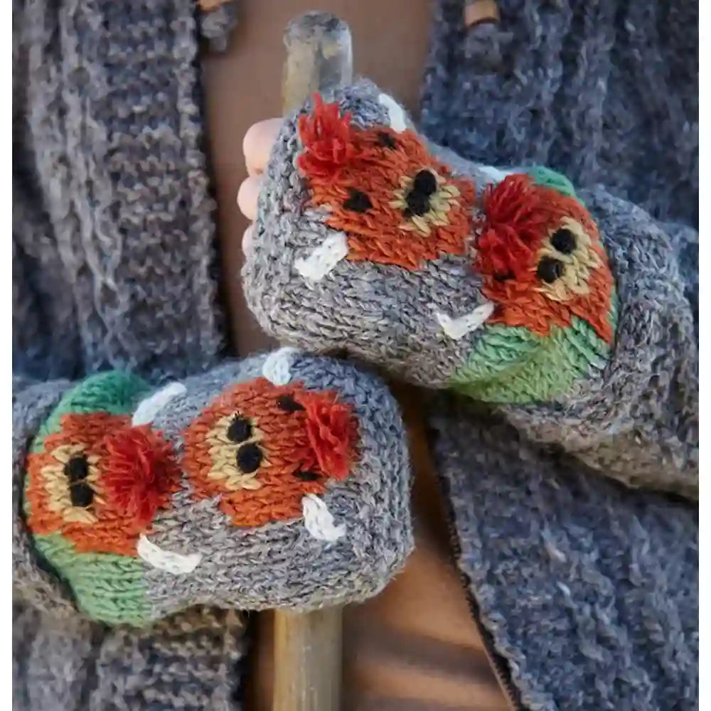 A close-up of a person wearing gray knit mittens with embroidered orange, red, green, and beige fox faces, holding a wooden stick outdoors.