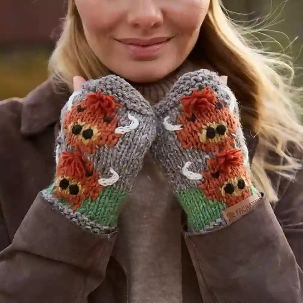 A close-up of a woman wearing gray knit fingerless gloves with a colorful embroidered design of orange foxes, green grass, and white deer on the back of the hands.