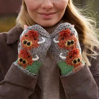 A close-up of a woman wearing gray knit fingerless gloves with a colorful embroidered design of orange foxes, green grass, and white deer on the back of the hands.