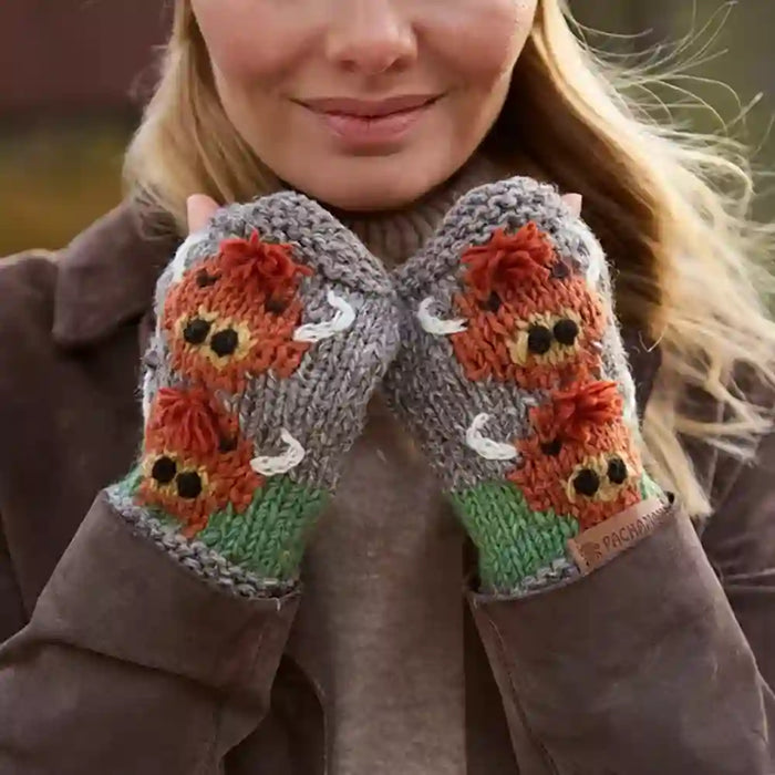 A close-up of a woman wearing gray knit fingerless gloves with a colorful embroidered design of orange foxes, green grass, and white deer on the back of the hands.