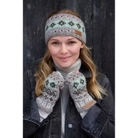 A woman wearing a knitted beanie and matching knit fingerless gloves with a Fair Isle pattern in sage green, grey, and brown, posed against a dark wooden background.