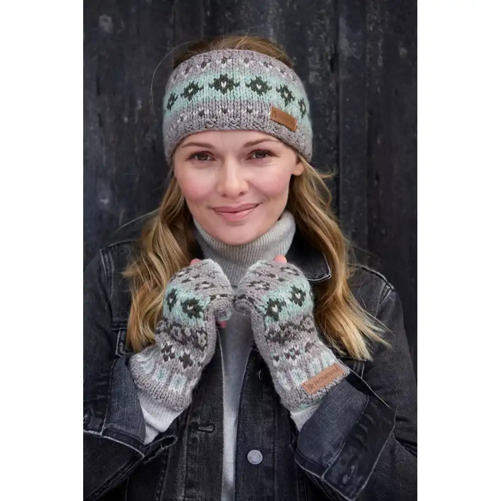 A woman wearing a knitted headband and matching knit fingerless gloves with a Fair Isle pattern in sage green, grey, and brown, posed against a dark wooden background.