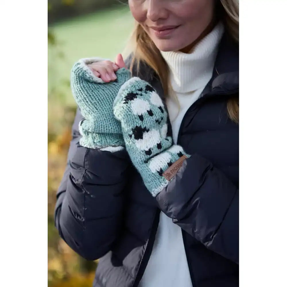 A close-up of a woman wearing knit fingerless gloves with a black and white sheep on a mint green pattern, paired with a white turtleneck and a dark navy puffer jacket.