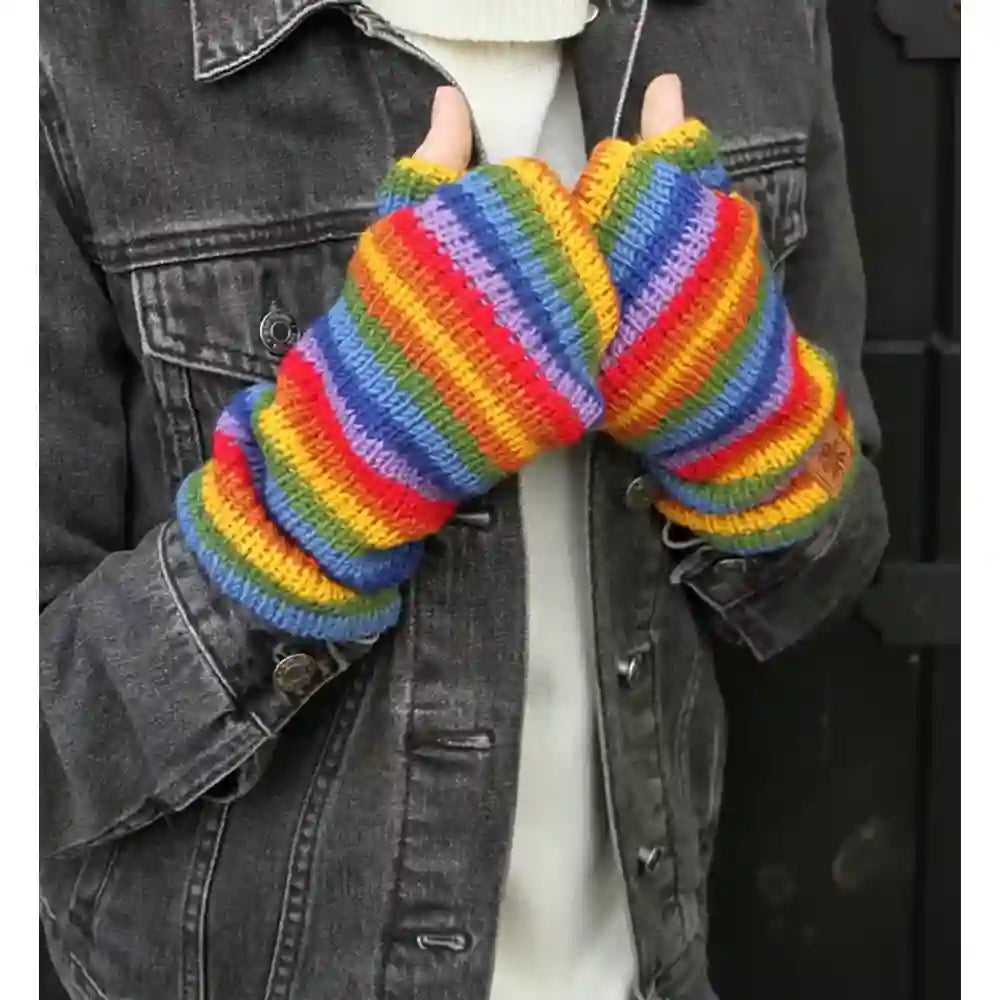 A person wearing a pair of chunky knit fingerless gloves with bold rainbow stripes in red, orange, yellow, green, blue, and purple, styled with a dark gray denim jacket.