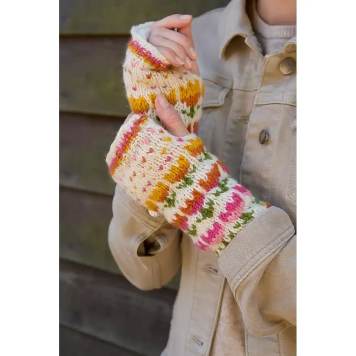 A person wearing cream knit fingerless gloves with multicolored floral patterns in pink, orange, yellow, and green, paired with a beige jacket.