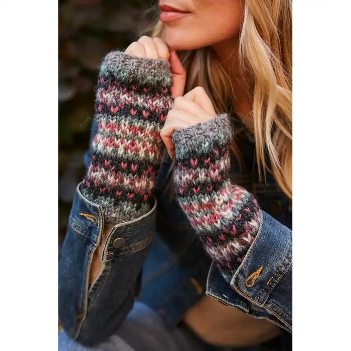 A close-up lifestyle shot of a woman wearing chunky knit fingerless wrist warmers with a multicolor Fair Isle pattern of pink, red, blue, and grey, styled under a denim jacket.