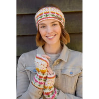 A smiling woman wearing a cream knit headband and matching fingerless gloves with multicolored Fair Isle-style flowers in pink, orange, yellow, green, and blue, posed against a dark wooden wall.