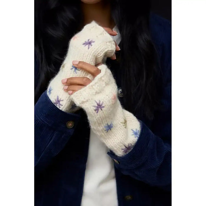 A person wearing cream knit fingerless gloves with small purple and blue flower patterns, shown against a dark navy corduroy jacket and white shirt.