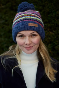 A woman wearing a colorful striped knit beanie with a pom-pom. The hat features horizontal stripes in blue, red, yellow, green, and white, with a small square logo patch on the front.