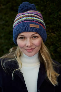 A woman wearing a colorful striped knit beanie with a pom-pom. The hat features horizontal stripes in blue, red, yellow, green, and white, with a small square logo patch on the front.