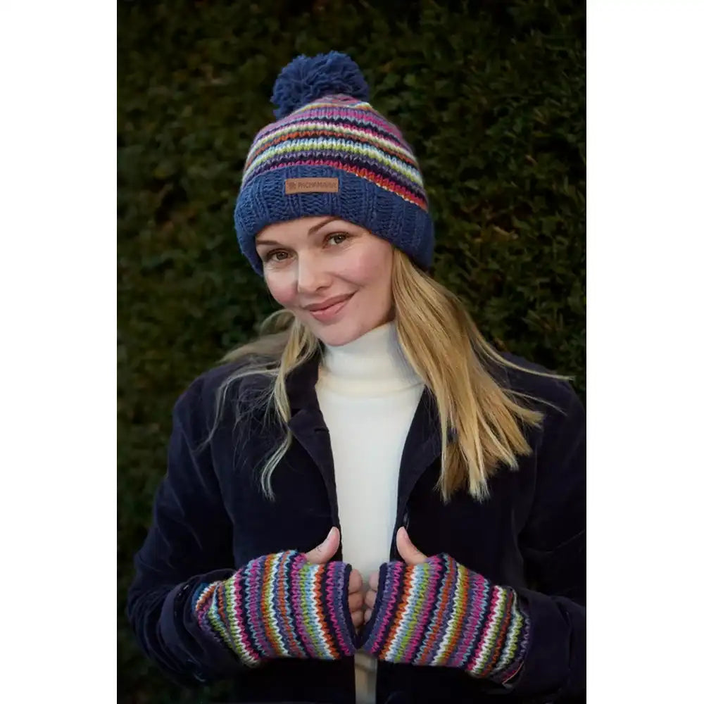 A woman wearing a colorful striped knit beanie with a pom-pom. The hat features horizontal stripes in blue, red, yellow, green, and white, with a small square logo patch on the front.  She is also wearing matchng handwarmers.