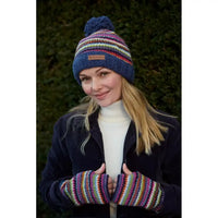 A woman wearing a colorful striped knit beanie with a pom-pom. The hat features horizontal stripes in blue, red, yellow, green, and white, with a small square logo patch on the front.  She is also wearing matchng handwarmers.