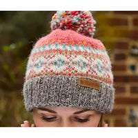 A close-up of a person wearing a knitted pom-pom beanie. The hat features a pink top section with a multicolor Fair Isle pattern, a gray ribbed cuff, and a small brown leather patch on the side.