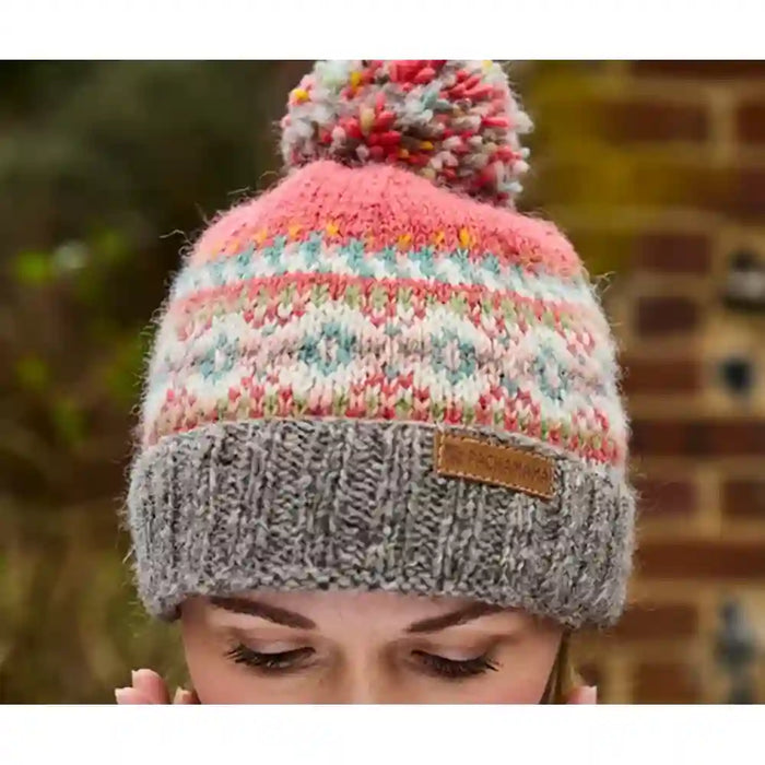 A close-up of a person wearing a knitted pom-pom beanie. The hat features a pink top section with a multicolor Fair Isle pattern, a gray ribbed cuff, and a small brown leather patch on the side.