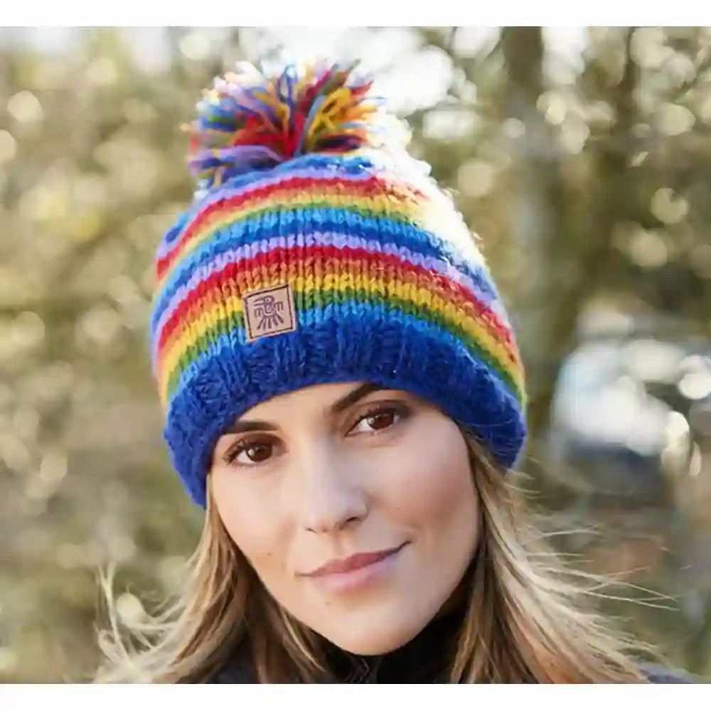 A woman wearing a colorful striped knit beanie with a pom-pom. The hat features horizontal stripes in blue, red, yellow, green, and white, with a small square logo patch on the front.