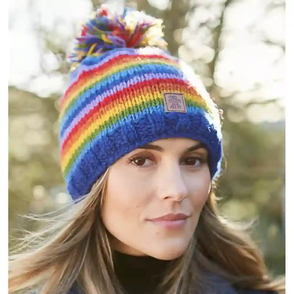 A woman wearing a vibrant rainbow-striped knit beanie with a matching pom-pom and a small Union Jack patch on the front, set against a blurred outdoor background.