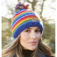 A woman wearing a vibrant rainbow-striped knit beanie with a matching pom-pom and a small Union Jack patch on the front, set against a blurred outdoor background.