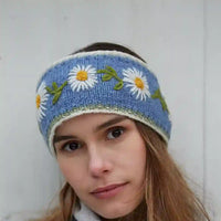 A woman wearing a light blue knit headband decorated with embroidered white daisies that have yellow centers and green leaves.