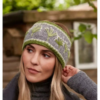 A woman wearing a gray knit headband with green and white leaf patterns, photographed outdoors with a blurred wooden structure and greenery in the background.