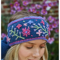 A woman wearing a navy blue knitted headband with colorful floral embroidery in pink, purple, and green, photographed outdoors with blurred flowers in the background.