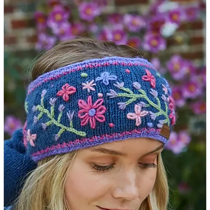 A woman wearing a navy blue knitted headband with colorful floral embroidery in pink, purple, and green, photographed outdoors with blurred flowers in the background.